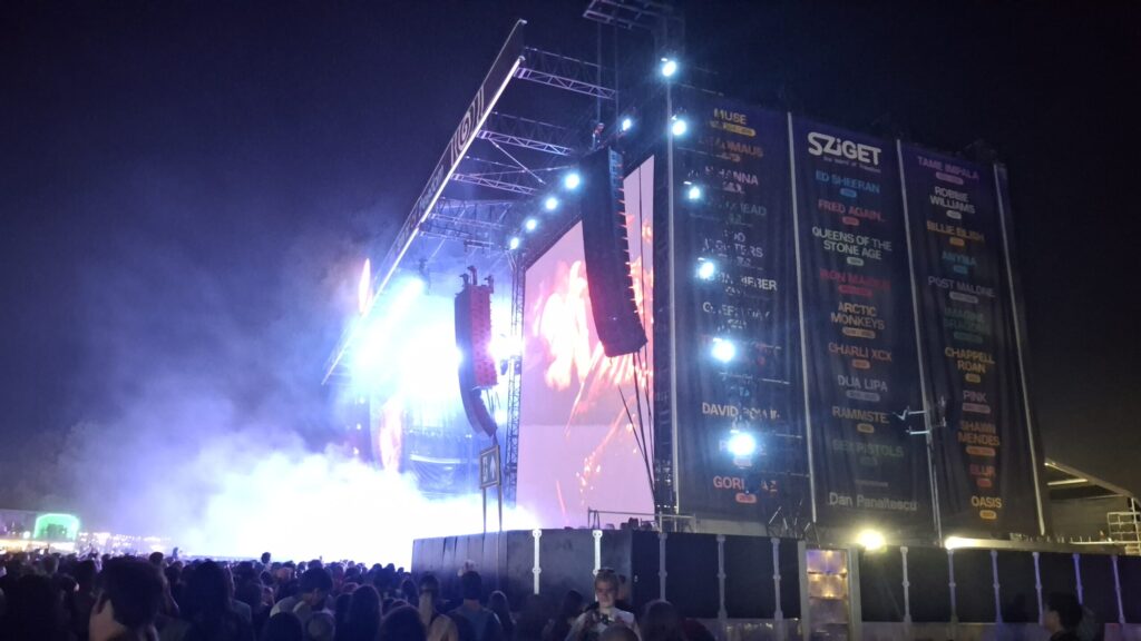 A nighttime shot of a large festival stage at the Sziget Festival. Spotlights and LED screens illuminate the stage, while a dense crowd gathers in front of it. Large banners featuring the Sziget logo and the names of various artists hang from the stage structure on the side.